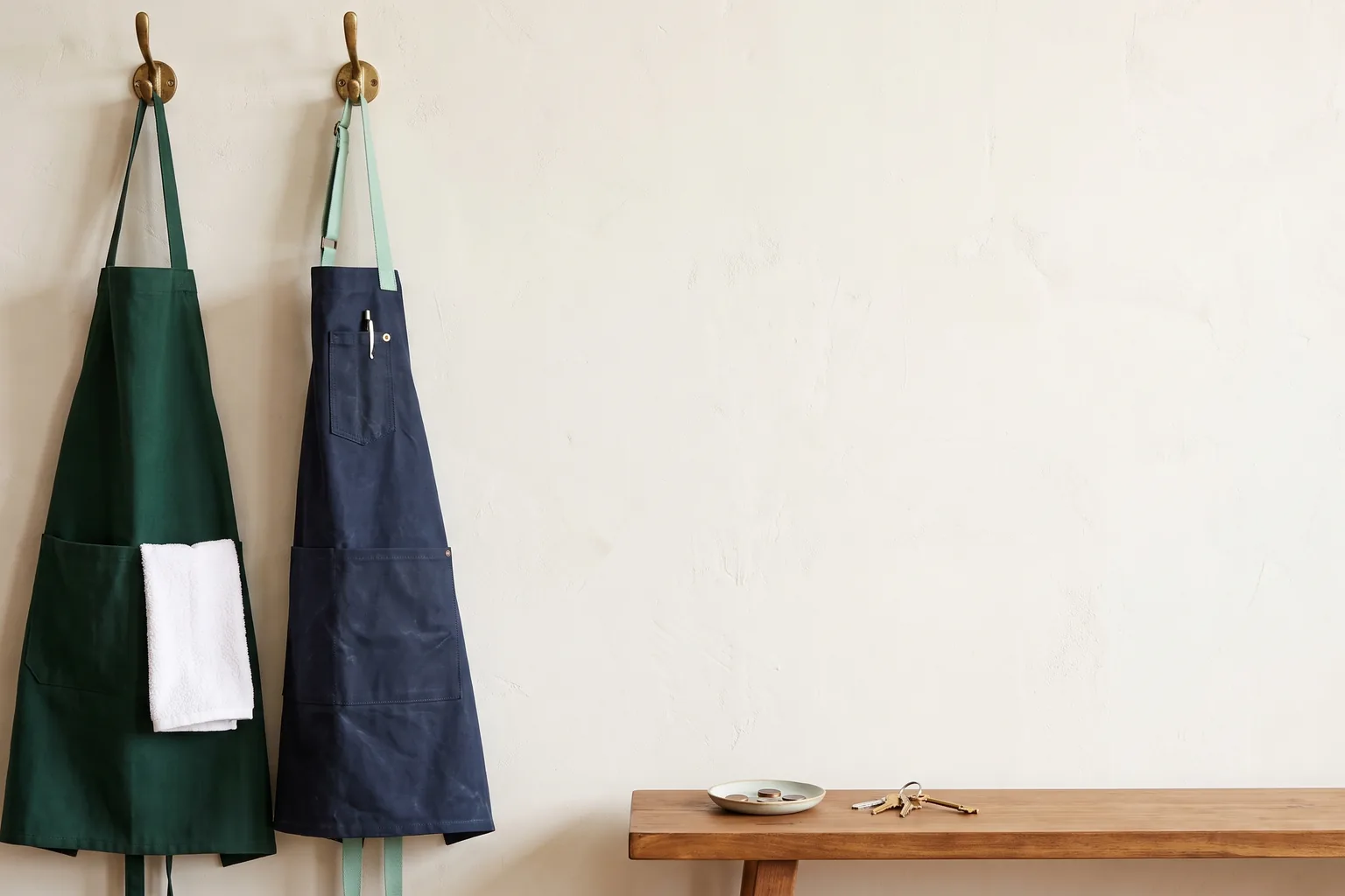 A green bib apron and a navy café apron hanging on brass hooks above a wooden bench holding a dish of coins and keys
