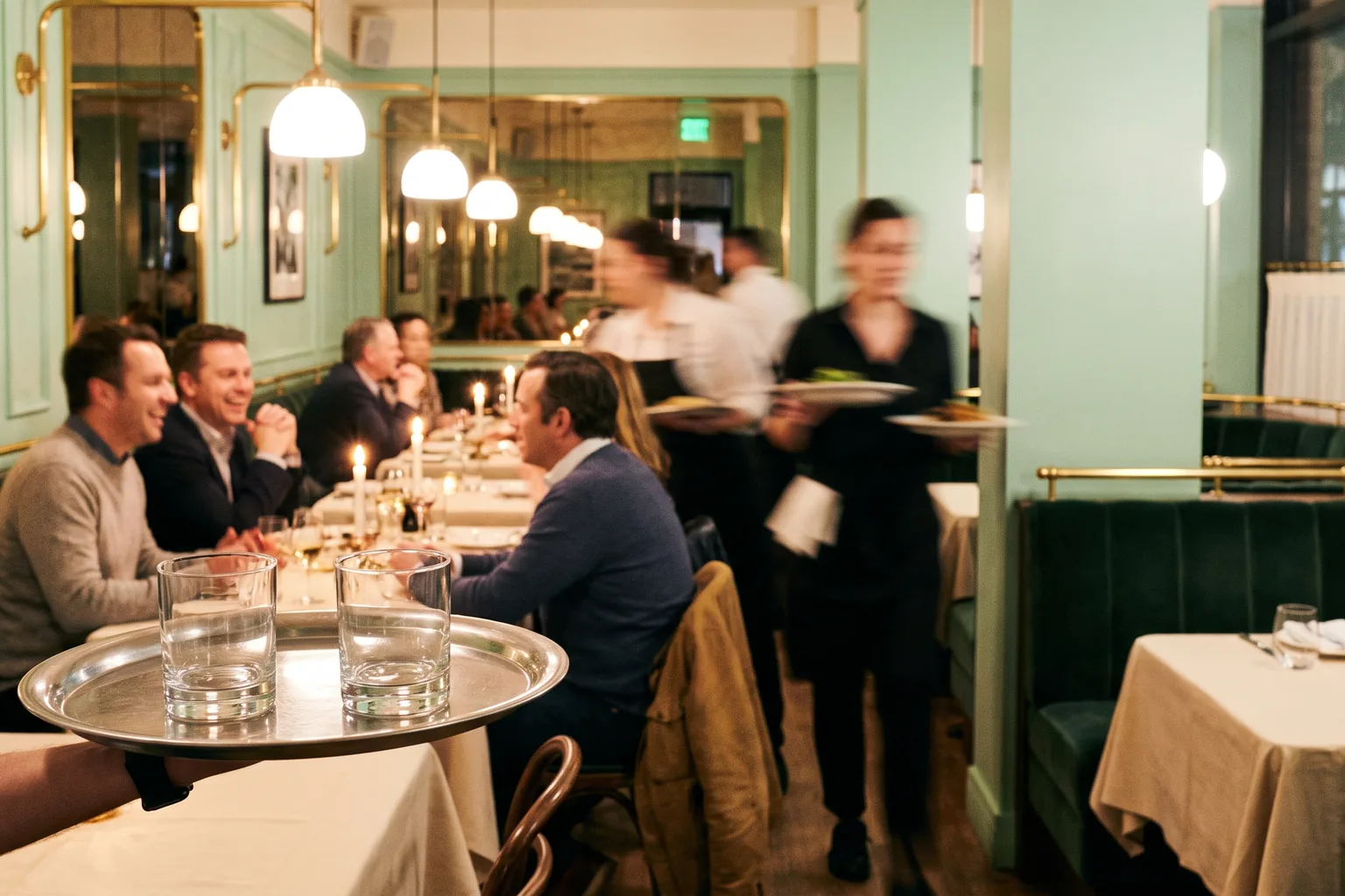 A busy restaurant dining room at night seen over a server's tray, with green banquettes, brass lamps and blurred staff in motion